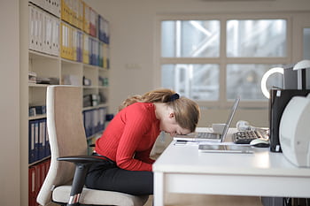 Tired woman in red sweater naps on office desk beside laptop, overwhelmed by remote work. HD wallpaper