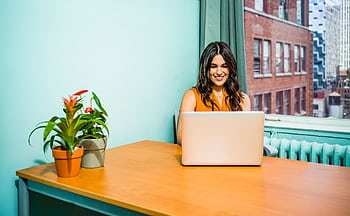 Confident woman in a bright office working on a laptop, surrounded by urban scenery and greenery. HD wallpaper