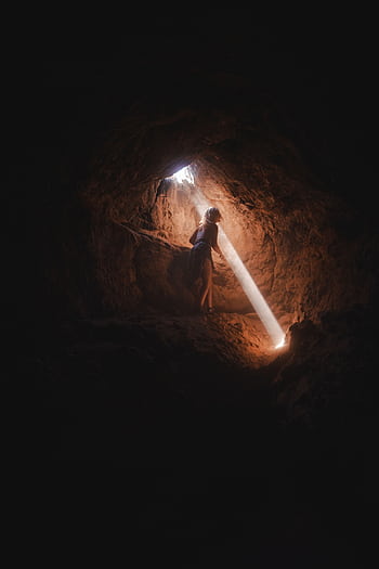 A lone adventurer explores a dark cave, illuminated by a dramatic sunbeam. HD phone wallpaper