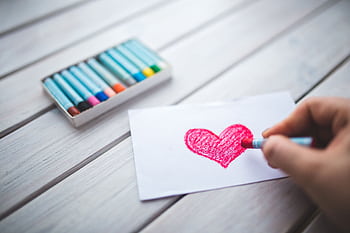 A close-up of a hand using pastel crayons to draw a red heart on paper. HD wallpaper