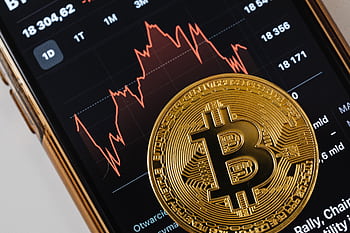 A close-up of a gold Bitcoin coin placed on a smart displaying a stock market chart. HD wallpaper