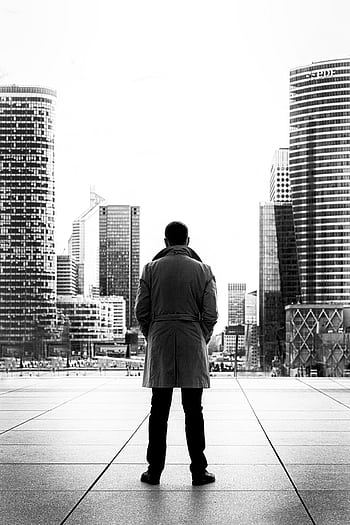 Silhouette of a man in a trench coat standing against a modern urban cityscape in black and white. HD phone wallpaper