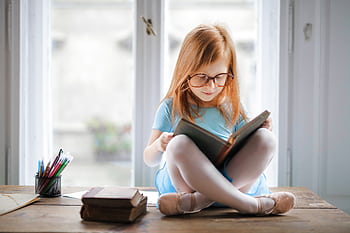 Adorable young girl wearing glasses, reading a book indoors by a window. Captures innocence and focus. HD wallpaper