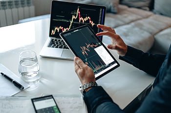 Close-up of a person using a tablet for online trading analysis with a laptop and glass of water nearby. HD wallpaper