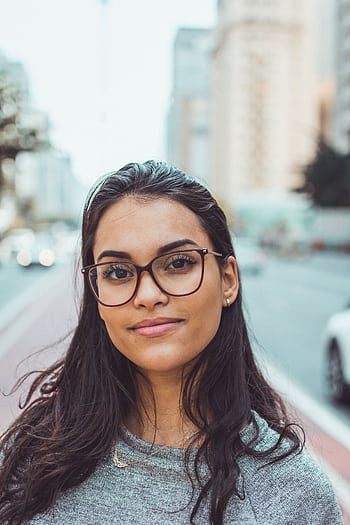 A confident woman wearing glasses smiles subtly while standing on an urban street with blurred background. HD phone wallpaper