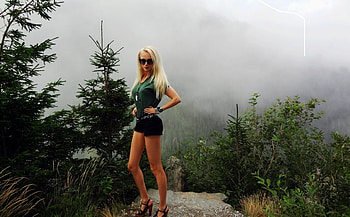 Valeria Lukyanova Barbie Doll look, standing on rock, blonde, hot black shorts, jewelry, evergreens, sunglasses, mountain in background, heels, forest green blouse, belt, misty view HD wallpaper