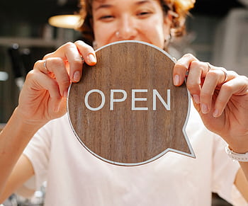 Happy woman holding an 'Open' sign at a café, inviting customers indoors. HD wallpaper