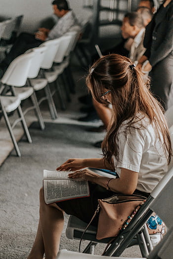 Focused woman reading a book in an academic setting with others in the background. HD phone wallpaper