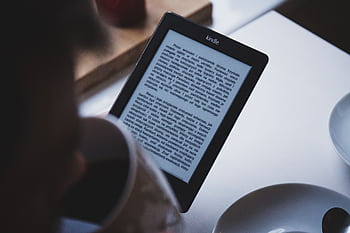A person reads on a Kindle e-reader while holding a cup of coffee, indoors. HD wallpaper