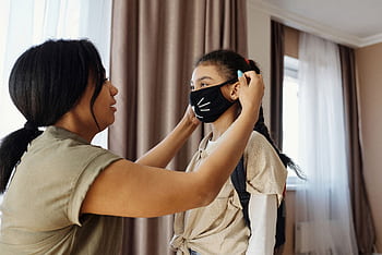 Mother assists daughter with wearing a face mask indoors for safety. HD wallpaper