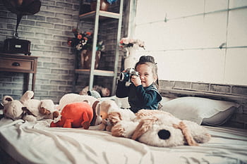 Smiling child with a camera surrounded by stuffed toys in a cozy bedroom setting. HD wallpaper