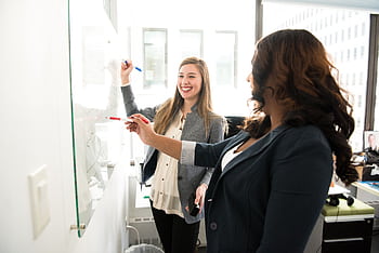 Two professional women discussing ideas on a whiteboard in a modern office setting. HD wallpaper