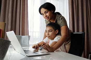 A mother helps her daughter with an online lesson on a laptop, showcasing family bonding and education at home. HD wallpaper