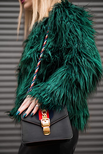 Stylish woman in a green faux fur jacket holding a fashionable handbag outdoors. HD phone wallpaper