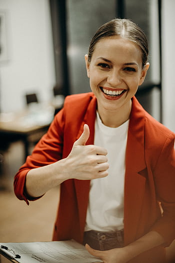 Confident businesswoman in a red blazer gives a thumbs up in a modern office setting. HD phone wallpaper