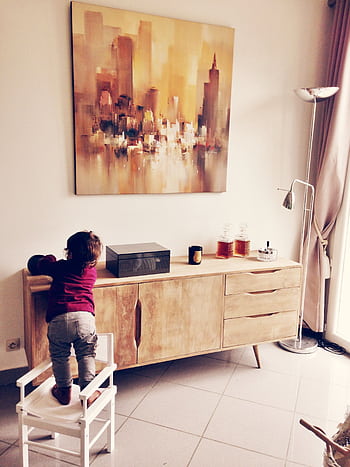 Child standing on chair reaching a wooden cabinet in a modern living room with abstract painting. HD phone wallpaper