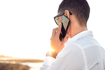Man outdoors in Morocco talking on smart, wearing sunglasses and white shirt, backlit by sunset. HD wallpaper