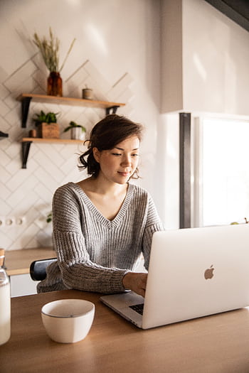 A woman in a sweater using a laptop at kitchen table, embracing remote work lifestyle. HD phone wallpaper
