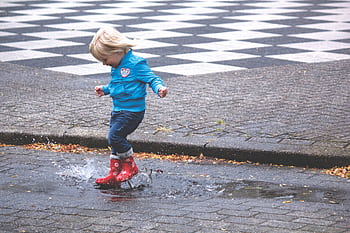 A young child joyfully splashes in a puddle wearing red boots on a rainy day. HD wallpaper