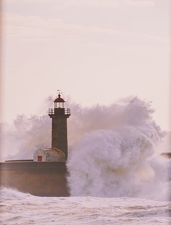 Powerful ocean waves crash against a solitary lighthouse at sunset, creating a dramatic and captivating scene. HD phone wallpaper