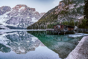 Serene winter landscape with snow-capped mountains and a lakeside cabin reflecting on a calm alpine lake. HD wallpaper