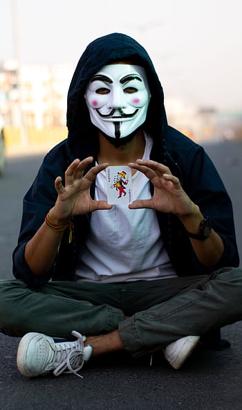 A person wearing an iconic mask sitting cross-legged and holding a joker card outdoors. HD phone wallpaper
