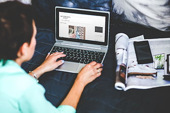 A woman on a bed browsing and blogging with a laptop and a magazine nearby. HD wallpaper