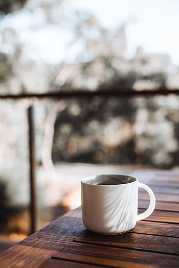 White coffee cup on a wooden table with blurred outdoor background, conveying a cozy and warm morning vibe. HD phone wallpaper