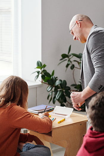 A teacher helps students during a classroom activity with books and notepads. HD phone wallpaper