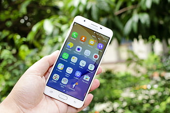 Close-up of a hand holding a smart displaying various app icons against a blurred outdoor background. HD wallpaper