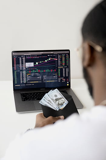 A person examines stock market data on a laptop while holding dollar bills, showcasing finance and trading concepts. HD phone wallpaper