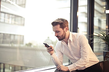 A businessman in a white shirt texts on his smart by a large glass window inside an office. HD wallpaper