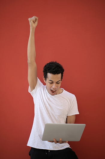 A young man in a white shirt celebrates success holding a laptop against a red background. HD phone wallpaper