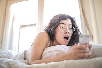 Young woman lying on bed, surprised by something on her smart, in a bright bedroom setting. HD wallpaper