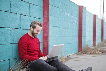 A man smiling and using a laptop outdoors against a vibrant blue wall, representing remote work. HD wallpaper