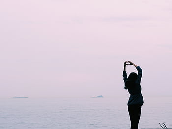 A silhouette of a woman making a heart shape with her hands by the calm sea during sunset. HD wallpaper
