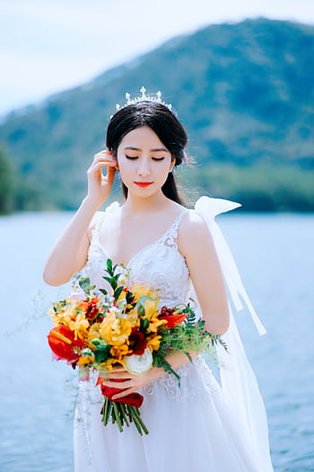 Bride in white wedding gown holding a colorful bouquet with a serene lake backdrop in Đà Lạt, Vietnam. HD phone wallpaper