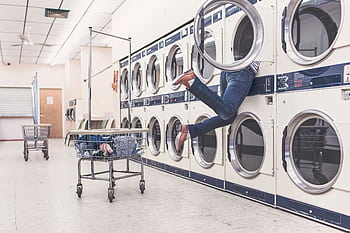 A funny scene in a laundromat with a person partially inside a dryer, highlighting everyday mishaps. HD wallpaper