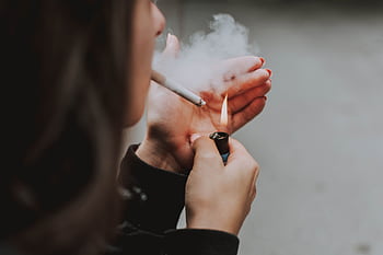 Close-up of a woman lighting a cigarette outdoors with a lighter, smoke visible. HD wallpaper