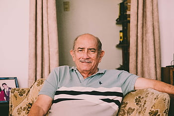 A cheerful elderly man enjoys a relaxed moment on a sofa in a warmly decorated living room. HD wallpaper