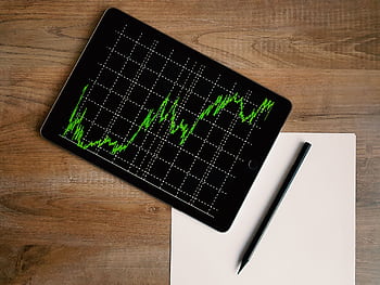 Flatlay of an iPad displaying stock market graph on a wooden desk with a pencil and paper. HD wallpaper