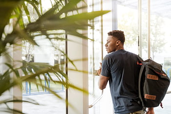 A college student with a backpack exits a campus building through glass doors. HD wallpaper