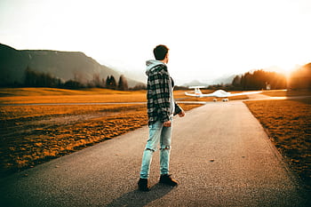 A man in casual attire stands on a road, observing a plane at sunset in Ohlstadt, Germany. HD wallpaper