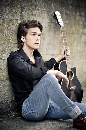 Casual portrait of a young male musician with an acoustic guitar sitting against a wall outdoors. HD phone wallpaper