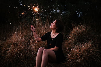 A smiling woman enjoying sparklers outdoors in a grassy field at night, expressing pure joy. HD wallpaper