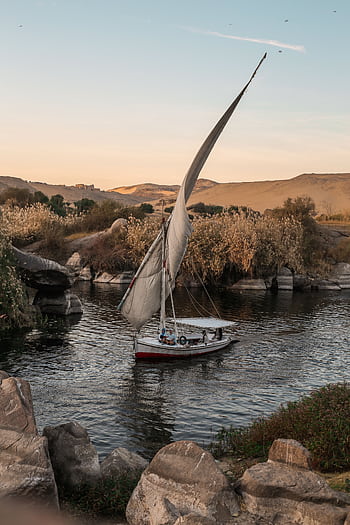 A serene sailboat glides along a river surrounded by rocks and hills during golden sunset light. HD phone wallpaper