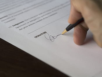A hand signs a formal contract with a pen on a wooden desk. HD wallpaper