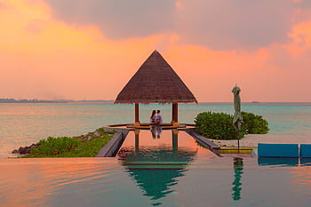 A couple enjoying a romantic moment by the sea and pool under a beach cabana during sunset. HD wallpaper