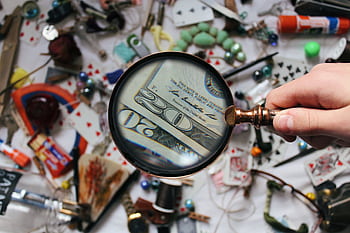A hand holding a magnifying glass focusing on a twenty dollar bill amidst various scattered objects. HD wallpaper