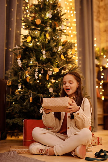 A joyful teenage girl opens a present by the Christmas tree, filled with excitement and seasonal cheer. HD phone wallpaper